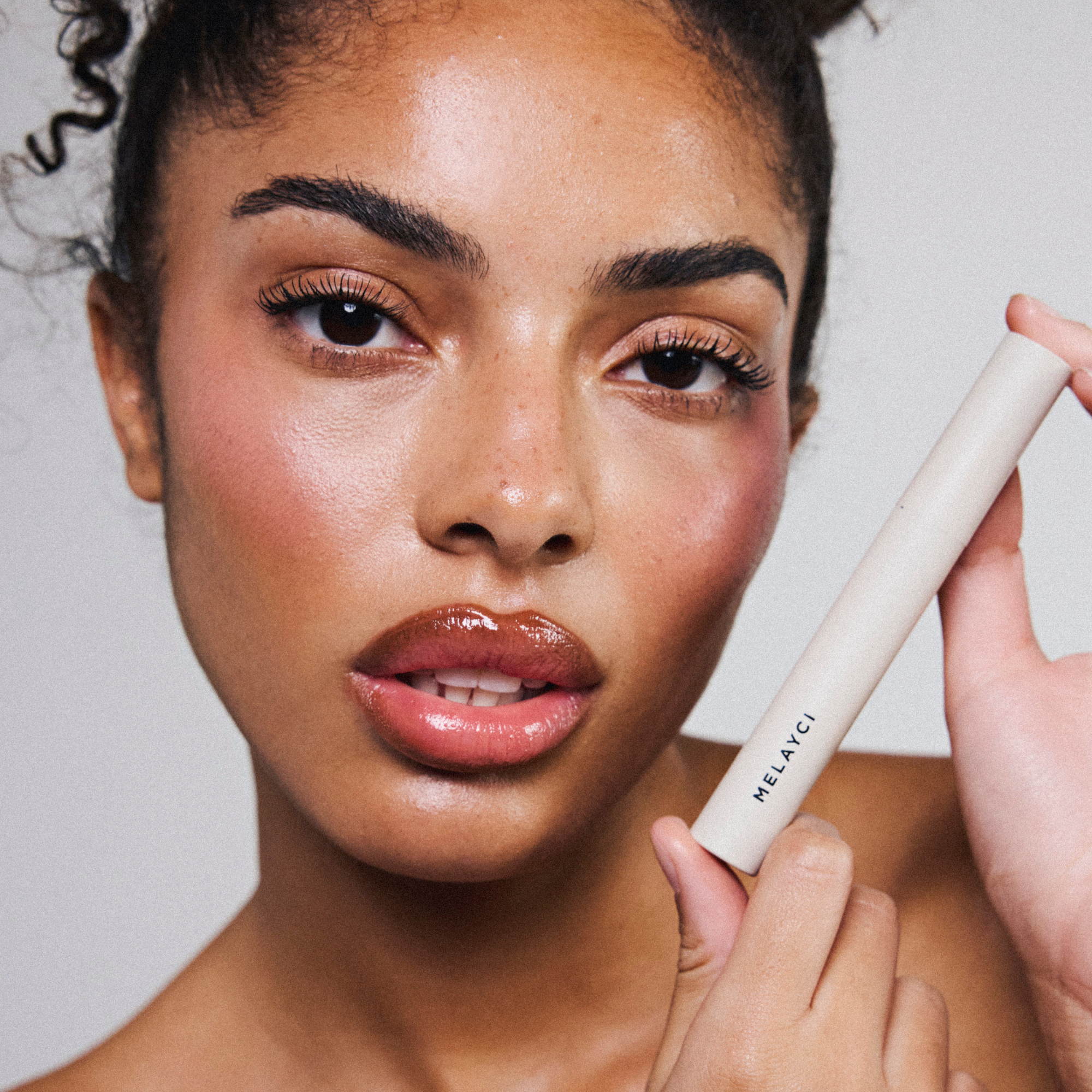 Modèle femme à la peau métissée portant le mascara, le gloss et le crayon à lèvres Melayci, tenant le mascara dans ses mains.
